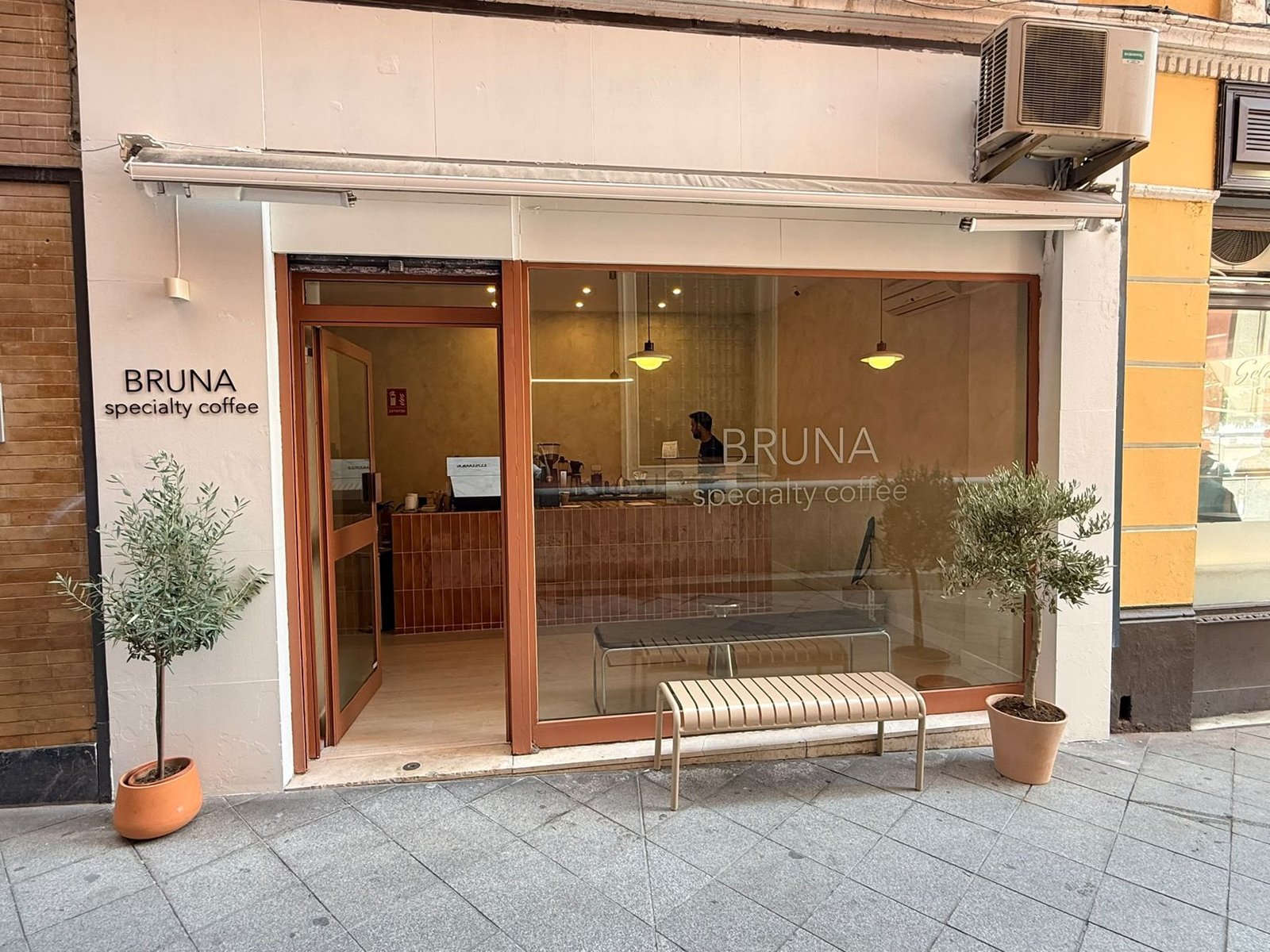 Fachada de Bruna Specialty Coffee, con marcos terracota, un banco y dos olivos en macetas